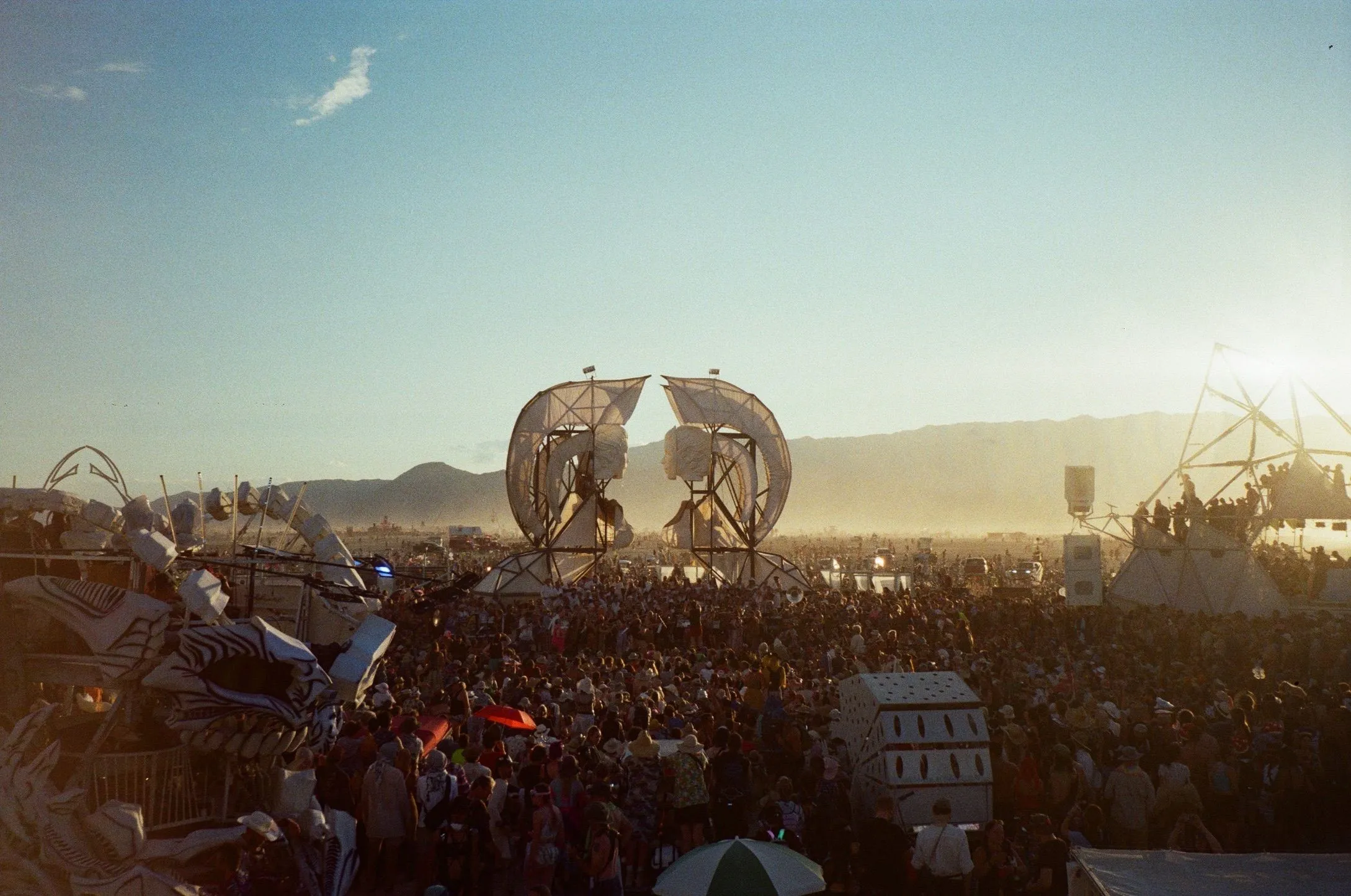 A dense crowd on dusty desert flats in low sun, gathered around two giant white sculpted faces, mountains behind