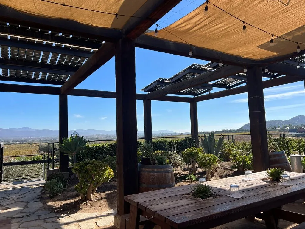 A shaded patio with a long wooden table and wine-barrel planters, looking out over vineyards to dry hills