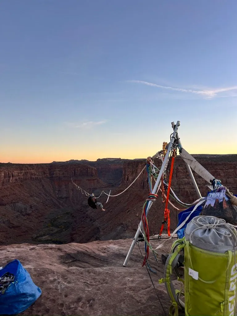 A person on a highline over a red-rock canyon at twilight, the line rigged from a metal A-frame on the rim